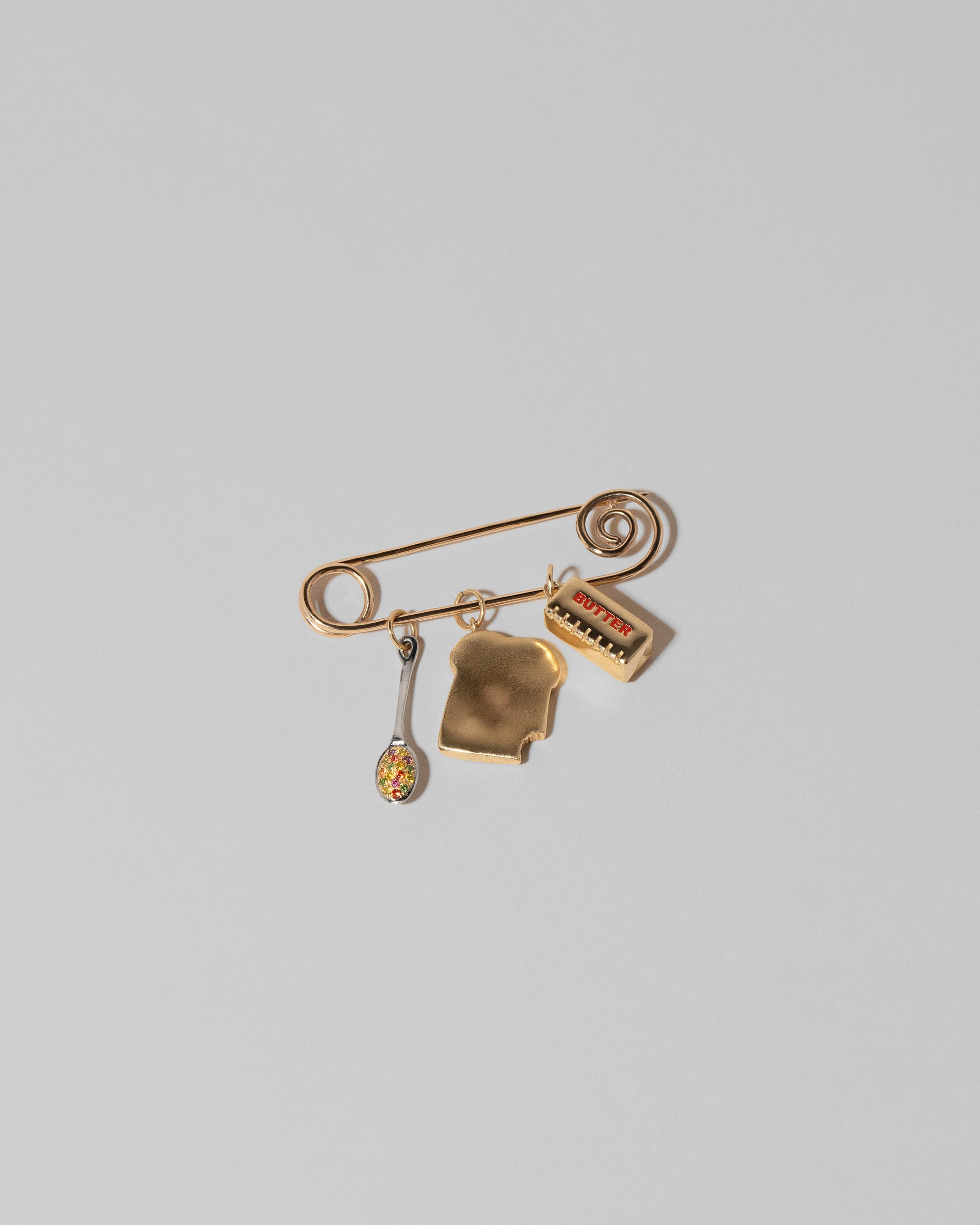 Detail view of the Spiral Pin Brooch with Cereal Spoon Charm, Bitten Toast Charm and Stick of Unsalted Butter Charm on light color background.